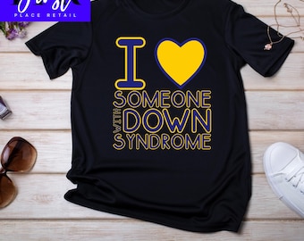 May include: Black t-shirt with the text "I heart someone with Down Syndrome" in yellow and blue. The heart symbol is yellow. The shirt is displayed on a light wood surface.