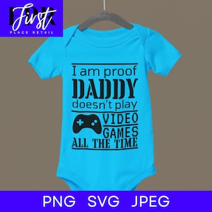 Puede incluir: Un body de bebé azul claro con texto negro que dice "Soy la prueba de que papá no juega a los videojuegos todo el tiempo". Se incluye una imagen en blanco y negro de un mando de videojuegos.