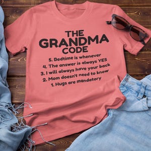 May include: A coral t-shirt with the words "THE GRANDMA CODE" in black, and a list of rules. The shirt is displayed with blue jeans, sunglasses, and a blue scarf on a wooden surface.