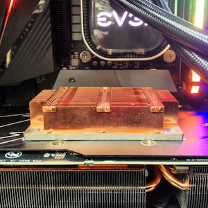 May include: Close-up of a copper heatsink with a black and silver heatsink cover. The heatsink is attached to a black graphics card with the text "GEFORCE" visible. The graphics card is part of a computer system with a water cooling system with black tubing and a clear water block with the text "EVGA" visible.