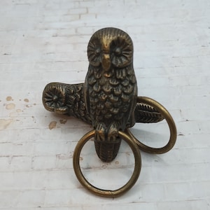 May include: A gold-toned owl figurine with detailed feathers and a ring attached to its feet. The owl is perched on a branch with another ring attached to the branch.