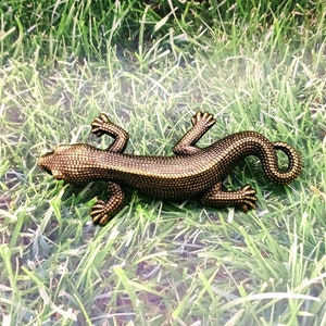 May include: A bronze-colored lizard figurine with a textured surface, lying on a bed of green grass.