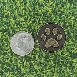 May include: A round, bronze-colored golf ball marker with a paw print design, next to a U.S. dime coin. The marker has a dark gray background with a bronze-colored paw print. The dime is silver and shows a profile of a person.