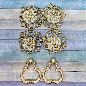 May include: Four decorative metal drawer pulls with ornate, square and circular filigree backplates in gold and white, featuring a central floral motif. Below these are two matching gold metal drop-style drawer handles with curved, symmetrical shapes.
