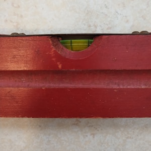 May include: A red wooden level with a green bubble level. The level is used to determine if a surface is horizontal or vertical.