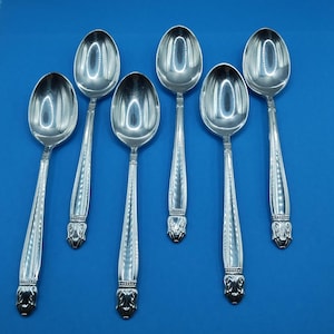 May include: Six silver spoons with oval bowls and ornate handles are arranged against a solid blue background. The spoons have a reflective surface, and the handles feature a decorative design at the base.