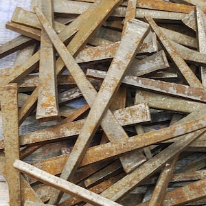 May include: A pile of weathered, rectangular metal pieces with a rusty, aged appearance. The metal pieces are various sizes and shapes, with a flat head on one end and a tapered point on the other. The pieces are scattered and overlapping.