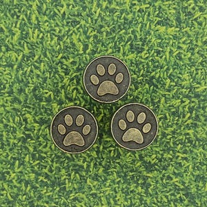 May include: Three bronze-colored paw print charms on a green background. Each charm has a dark gray border and a detailed paw print design in a lighter bronze hue. The charms are likely for jewelry or crafts.