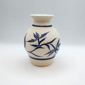 May include: A cream-colored ceramic vase with a rounded body and a flared rim. The vase features hand-painted dark blue floral motifs encircling the center, complemented by two horizontal dark blue bands near the top and bottom of the vessel.