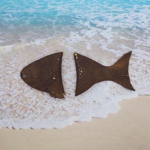 May include: Two brown fish sculptures with gold accents, placed on a sandy beach with white foamy waves.