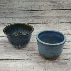 May include: Two ceramic bowls, one with a dark blue and green glaze and the other with a light blue glaze. Both bowls have a rustic, handmade look.