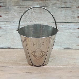 May include: A small, silver-colored metal bucket with a curved handle. The word "FIRE" is etched on the front, along with a fire department emblem. The bucket is likely used for decorative purposes or as a small container.