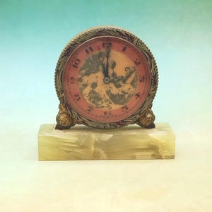 May include: A round analog clock with a metallic frame featuring an intricate embossed pattern. The clock face has a pink border with black numerals and a central illustration of figures. It rests on a rectangular, light-colored stone base.