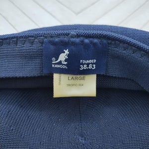 Op de afbeelding: Een donkerblauwe Kangol-pet van getextureerde gebreide stof. De binnenkant toont een marineblauw label met een wit kangoeroelogo en de tekst "KANGOL FOUNDED 38.83". Een cr&egrave;mekleurig label geeft de maat "LARGE" en "TROPIC 504" aan.