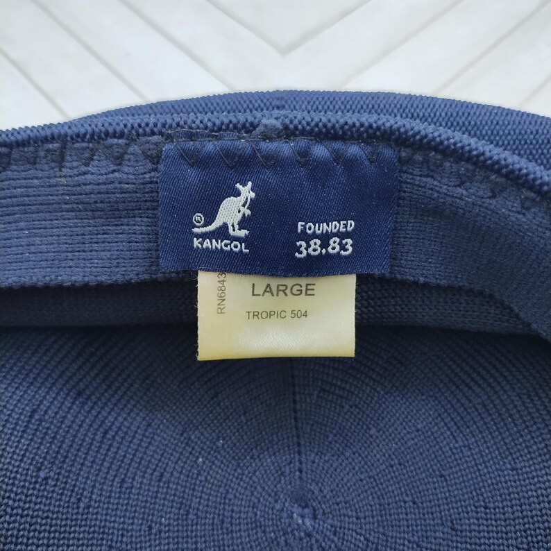 Op de afbeelding: Een donkerblauwe Kangol-pet van getextureerde gebreide stof. De binnenkant toont een marineblauw label met een wit kangoeroelogo en de tekst "KANGOL FOUNDED 38.83". Een cr&egrave;mekleurig label geeft de maat "LARGE" en "TROPIC 504" aan.