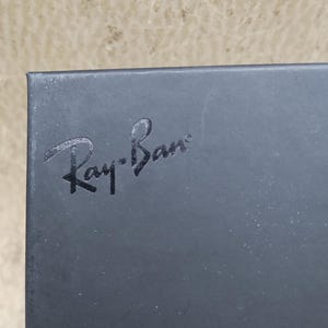 Puede incluir: Primer plano de una caja gris oscuro con el texto en relieve "Ray-Ban" en una fuente estilizada. La caja tiene una superficie ligeramente texturizada y un acabado mate. El fondo es de un color neutro claro y borroso.