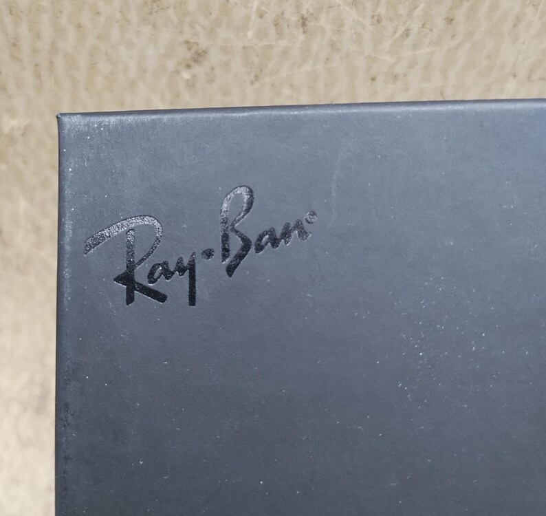 Puede incluir: Primer plano de una caja gris oscuro con el texto en relieve "Ray-Ban" en una fuente estilizada. La caja tiene una superficie ligeramente texturizada y un acabado mate. El fondo es de un color neutro claro y borroso.