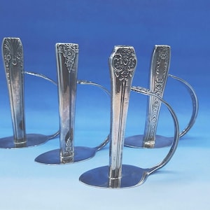May include: Four silver candle holders crafted from spoons. Each holder features a spoon bowl base and a curved handle that supports the candle. The spoon handles are upright, with decorative floral patterns. The background is a gradient of blue.