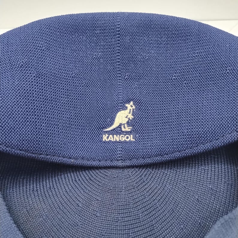 Op de afbeelding: Een donkerblauwe Kangol-pet van getextureerde gebreide stof met een opvallend geborduurd logo aan de voorzijde. Het logo toont een wit kangoeroesilhouet boven het woord KANGOL, gecentreerd op de afgeronde bovenkant van de pet.