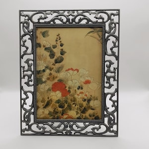 May include: A rectangular picture frame with a dark gray, ornate, openwork metal border featuring scrolling patterns. The frame holds a print depicting a botanical scene with white and red flowers, green leaves, and stems against a beige background.