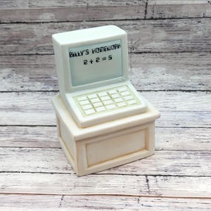 May include: A cream-colored, ceramic computer figurine with the text "BILLY'S HOMEWORK" and "2 + 2 = 5" on the screen. The figurine has a keyboard and sits atop a small, rectangular base. The background is a light wood surface.