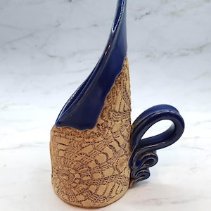 May include: A handmade ceramic mug with a unique design. The mug features a textured, tan-colored body with a detailed pattern and a deep blue, curved rim and handle. The handle has a decorative, swirling design.
