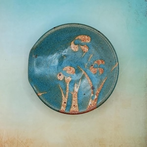 May include: A handmade ceramic spoon rest in a teal glaze with a floral design. The spoon rest is round with a small lip for resting utensils. The floral design features brown and white stylized flowers and stems.