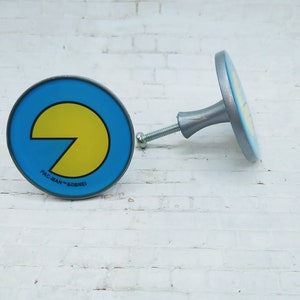 May include: Two Pac-Man themed cabinet knobs. The knobs are round and have a silver base. The front of the knobs are blue with a yellow Pac-Man graphic and the text "PAC-MAN™ & ©BANDAI".