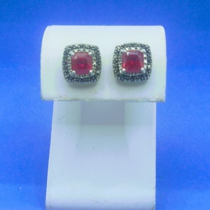 May include: A pair of silver earrings with a square red gemstone surrounded by smaller clear gemstones.