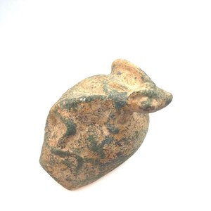 May include: A small, weathered, tan and green stone figurine shaped like a crouching animal. The object features a textured, uneven surface with visible dark green patina-like markings and a distinct head with pointed ears extending from the body.