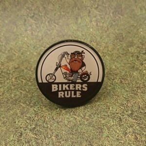 May include: A round black and white pin with a cartoon biker on a motorcycle. The text "Bikers Rule" is printed in black on a white background.