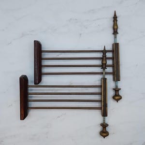 May include: Two antique towel racks. Each rack features a dark wood frame with horizontal metal bars. One rack has a decorative brass element with a threaded rod and finial. The other rack is missing the decorative element.