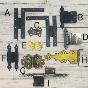 May include: Assortment of ten antique hinges in various styles and finishes, including black, silver, and brass. Some hinges have ornate designs, while others are more simple. The hinges are arranged on a wooden surface.