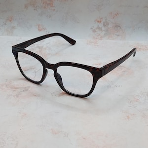 May include: A pair of eyeglasses with a tortoiseshell pattern. The frames are dark brown with a speckled design, and the lenses are clear. The glasses have a classic, rectangular shape with slightly rounded edges.