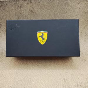 Puede incluir: Una caja rectangular negra con el logotipo de Ray-Ban en la esquina superior izquierda. El logotipo de Ferrari, un caballo rampante amarillo sobre un escudo amarillo con rayas verdes, blancas y rojas, est&aacute; centrado en la caja.