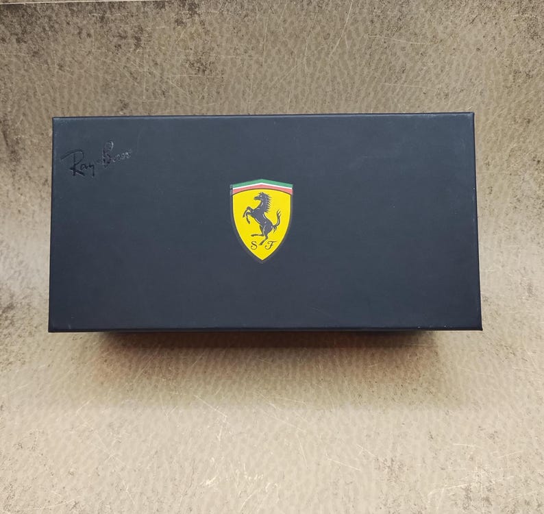 Puede incluir: Una caja rectangular negra con el logotipo de Ray-Ban en la esquina superior izquierda. El logotipo de Ferrari, un caballo rampante amarillo sobre un escudo amarillo con rayas verdes, blancas y rojas, est&aacute; centrado en la caja.