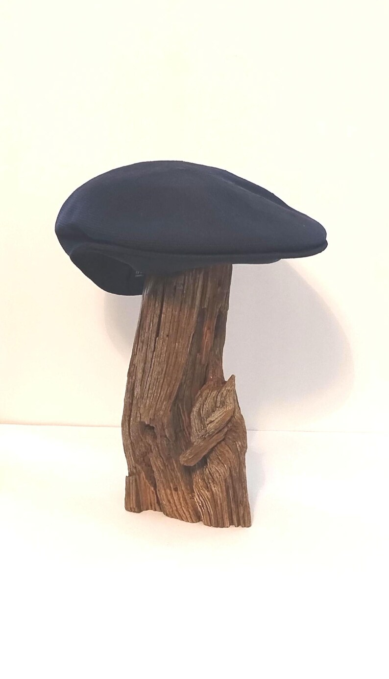 Op de afbeelding: Een effen donker marineblauwe platte pet met een afgeronde, gestructureerde bol en een korte, stijve klep. De pet wordt gepresenteerd op een stuk verweerd, getextureerd bruin drijfhout dat dient als standaard om de gladde stof te tonen.