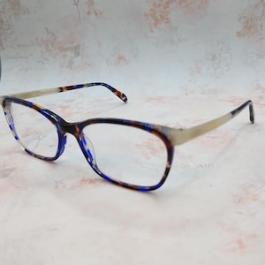 May include: Eyeglasses with a blue, brown, and white patterned frame and clear lenses. The temples are a light silver color. The glasses are set against a light pink and white background.