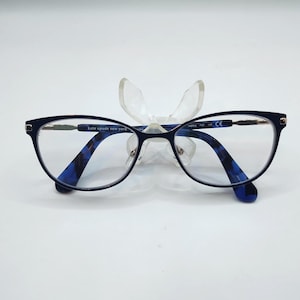 May include: Kate Spade New York eyeglasses featuring a dark navy blue front frame and temples with a blue and black tortoiseshell pattern on the inner side. The thin metal hinges connect the front to the temples, and the brand name is printed.