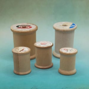 May include: Five vintage wooden spools of thread, varying in size, are arranged on a teal surface. The spools have light brown wooden bodies and circular tops with faded paper labels. Some labels show partial designs.