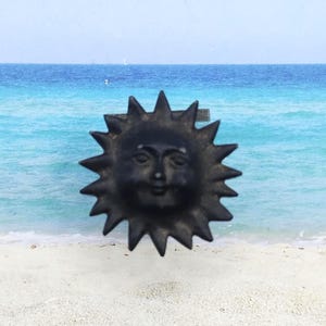 May include: A black sun-shaped decorative knob with a smiling face and spiky rays. The knob is made of a dark material and is set against a backdrop of a beach and ocean. The knob is likely for a cabinet or drawer.