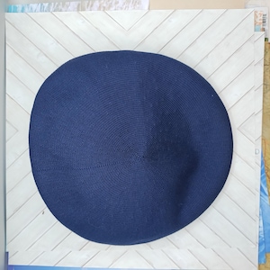 Op de afbeelding: Een cirkelvormige, donkerblauwe baret met een fijne, strak gebreide textuur. De hoed heeft een gladde, ronde kroon met een subtiel centraal punt en een consistent, uniform weefpatroon over het gehele oppervlak van de stof.