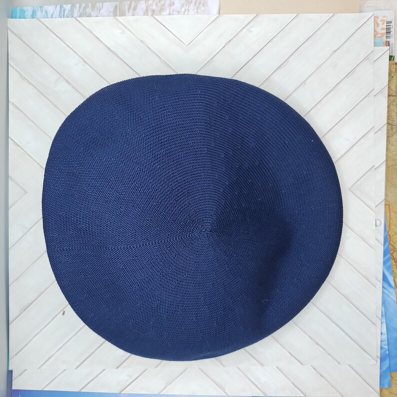 Op de afbeelding: Een cirkelvormige, donkerblauwe baret met een fijne, strak gebreide textuur. De hoed heeft een gladde, ronde kroon met een subtiel centraal punt en een consistent, uniform weefpatroon over het gehele oppervlak van de stof.