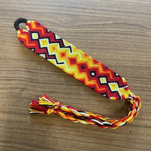 May include: A handmade friendship bracelet with a vibrant geometric pattern in red, yellow, orange, black, and white. The bracelet features a loop closure and braided ties with matching colors. The bracelet is laying on a wooden surface.