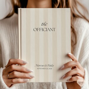 May include: A hardcover book with a cream and beige vertical striped cover. The front cover features the black text "the" and "OFFICIANT" in the center, with "Marcus & Haley" and "SEPTEMBER 26, 2026" printed in black near the bottom edge.