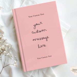 Puede incluir: Un diario de tapa dura rosa claro con el texto "Your Custom Text" en la parte superior e inferior, y "your custom message here" en el centro. El diario est&aacute; sobre una tela blanca con peque&ntilde;as flores blancas.