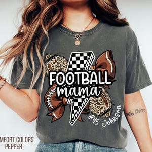May include: Dark grey Comfort Colors t-shirt with a football-themed graphic. The graphic includes a football, a bow, and the words "Football Mama". The shirt also features a lightning bolt and the name "Johnson".