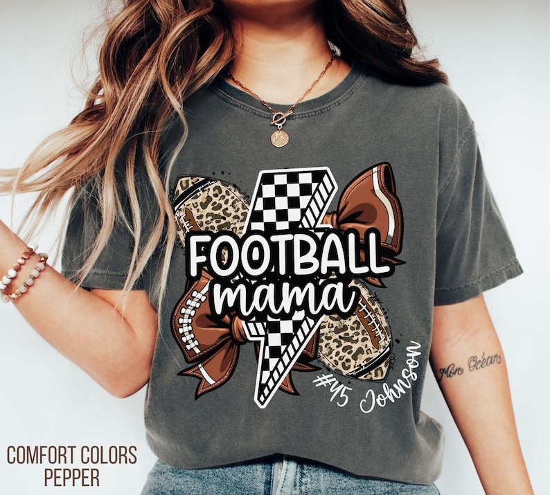 May include: Dark grey Comfort Colors t-shirt with a football-themed graphic. The graphic includes a football, a bow, and the words "Football Mama". The shirt also features a lightning bolt and the name "Johnson".