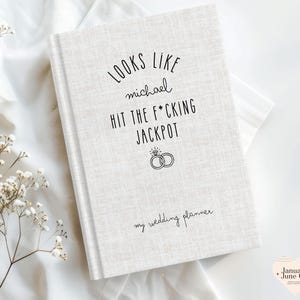 K&ouml;nnte beinhalten: Ein cremefarbener Hochzeitsplaner mit dem Text "Looks like Michael hit the f*cking jackpot" in schwarzer Schrift. Der Planer zeigt eine Ringgrafik und die Worte "my wedding planner" unten.