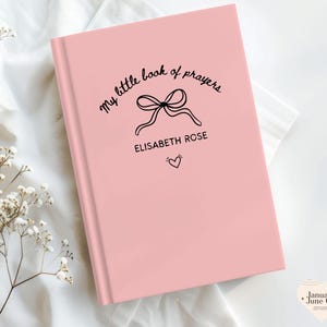 May include: A pink book with the text "my little book of prayers" in a handwritten font. Below the text is a bow illustration, the name "ELISABETH ROSE", and a small heart. The book is on a white surface.